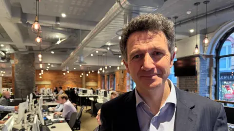 Dan Channer smiles for the camera inside his estate agent office. Rows of desks can be seen behind him with several people working at them. The large office has a modern feel with filament lights and exposed brick