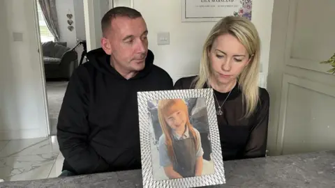 Lila's parents are looking at photograph of Lila in a silver frame. They are both sitting on stools in their kitchen. 