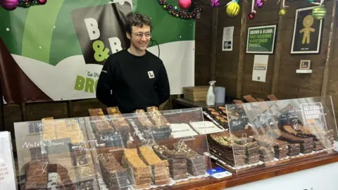 A man with short brown hair and glasses stands behind a stall selling chocolate brownies and blondies.