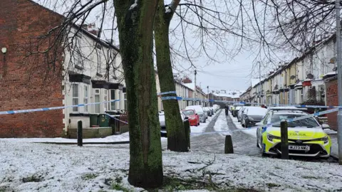 Police tape cordons off trees and end of a snowy road where police car is parked up at the end of a terrace of houses and parked cars