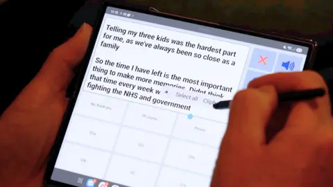 Marc Powell is pictured operating a handheld computer tablet. He has typed: "Telling my three kids was the hardest part" onto the screen.