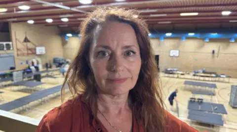 Bridget Kendrick stands on a balcony overlooking a sports hall. The lights are on behind her and she is wearing an orange Liberal Democrat badge.