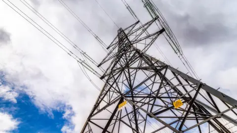A view looking up to the top of an electricity tower. The tower is a lattice work of metal beams. Six "arms" near the top are attached to power cables.