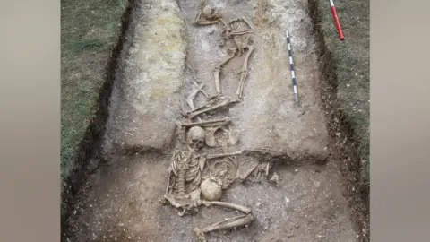 Cambridge Archaeological Unit/David Matzliach An archaeological excavation showing human remains at Wandlebury, Cambridgeshire. In the foreground is much of a skeleton on its back, with one arm above its head and another flung out to the right. Its right leg is bent up. There are other skulls and human remains behind it, including a largely intact skeleton.
