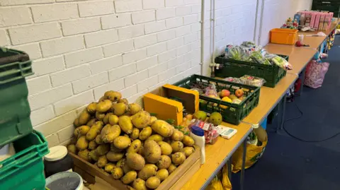 Boxes of food arranged on tables along a wall at Moorclose Community Centre in Workington. There is a box full of potatoes which are sprouting, lettuce, fruit and boxes of food.