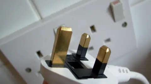 A close-up of a white three-point plug in front of a white wall socket.