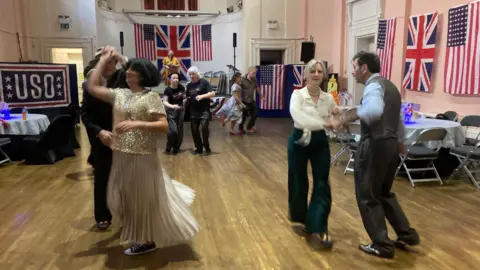 People dancing - holding hands - on  wooden floor, with American and British flags draped on the walls