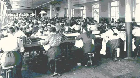 DCSDC Museum Services A group of women sit working in a shirt factory in the 1920s. There are several rows of women.