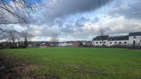 The old school playing field with houses visible beyond the hedgerow that borders the site