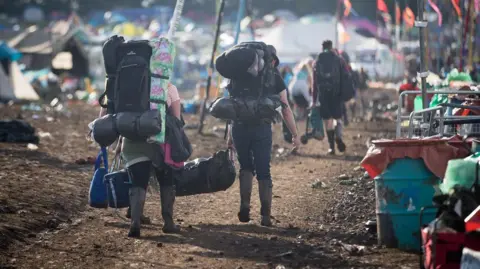 Getty Images People wearing backpacks leaving the Glastonbury Festival site. They are walking alongside a row of bins