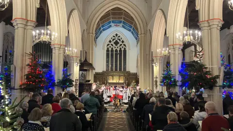 Jon Wright/BBC A picture taken from the back of a church, with a choir at the far end. The pews are full of people and there are Christmas trees around the pews.