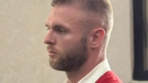 Thomas Paker sitting in court in Bali. He has short, light brown hair and a beard. He is wearing a white shirt and a red waistcoat, which defendants wear in Indonesian court.