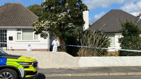 BBC Two white houses with brown roofs are surrounding with blue and white police tape as forensics in white suits examine the exterior. There is a blue, yellow and grey police car park on the street in front of the house.
