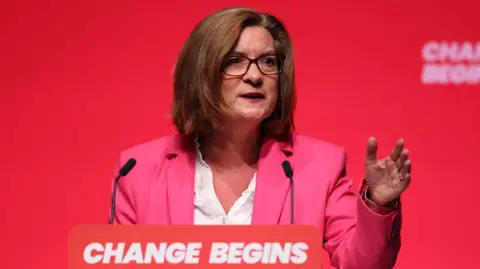 Eluned Morgan speaking at Labour conference in front of a banner that says "Change Begins". She is wearing a pink suit with a white shirt.