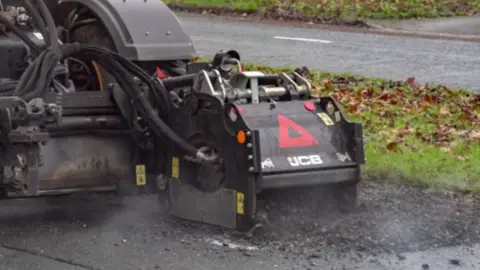 Leeds City Council The image shows a piece of heavy road‑repair machinery in operation on a residential street. The equipment is a black JCB-branded attachment connected to a larger vehicle outside the frame. It is positioned over damaged tarmac and is breaking up the surface, with loose debris and dust visible around the contact point. Several hydraulic hoses run from the attachment toward the vehicle. Behind the machinery, the road, a grass verge, and fallen leaves are visible.