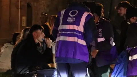 A group of volunteers in purple reflective jackets, with Street Angels Chester logos in white lettering, speak to a group of younger people sitting on a bench on a street at night time. A young man is drinking from a bottle of water.