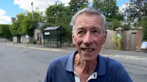 A man with grey hair, wearing a blue polo shirt, stands in front of a road with a bus stop behind him. He is pictured from the shoulders up.