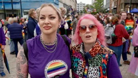A woman with blonde hair, tattoos and a purple tshirt alongside a woman with pink hair, sunglasses, and a multicoloured leopard-print top