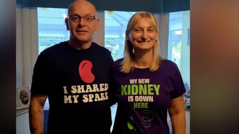 Allison and Darren Clay On the left, there is a man who is bald. He is wearing a black t-shirt with a red kidney shape, and the words 'I shared my spare' underneath. To his right, there is a woman with blonder hair. She is wearing a purple t-shirt with white text saying 'my new kidney is down here.' The letters in 'kidney' and 'here' are both in green and there is an arrow pointing to her kidney.