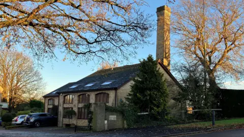 Current photo of the waterworks tower at Tuckton. A tall stone chimney can be seen behind a more modern two-storey brick house, which has four windows in its roof. Two cars are parked on the driveway. It is surrounded by trees which are bare from leaves.