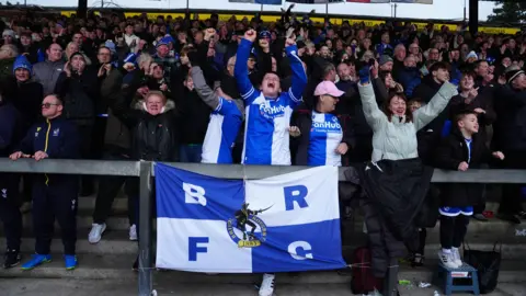 PA Media Bristol Rovers fans celebrate a goal in their game against Peterborough United at the Memorial Stadium. They are on a terrace and at the front is a large blue and white flag with "BRFC" in large letters and the club's crest on it