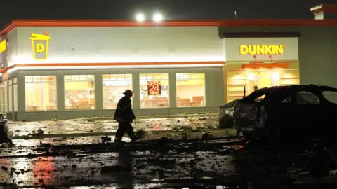 Getty Fire Photos walks down the road full of rubbish outside the Donuts' Dunkin branch. The burnt vehicle can be seen before him