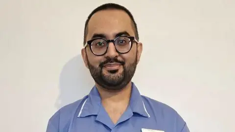 University of Leeds A male nurse stood in front of plain white wall, wearing a blue NHS nurses uniform. The nurse is wearing a pair of black framed glasses.