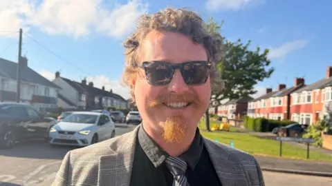 Jasmine Lowe/BBC A head and shoulders shot of a man standing in a residential street. He has short, brown curly hair and a ginger goatee and moustache. He is smiling at the camera and wearing a black shirt, a black and grey striped tie and a checked grey blazer.
