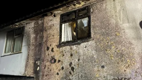 A picture of rendered houses with a window that's been shattered and blackened by fire. Smoke damage is apparent on the walls of the terraced properties.