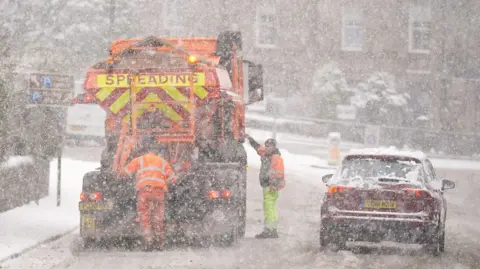 A gritter stopped on the side of the road in the snow with two workers in hi-vis around the vehicle. There is a dark red car passing by the side of the gritter.