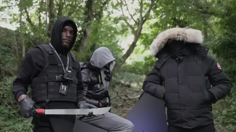 BBC Faron Paul who runs his own knife amnesty company holds a large knife. He is standing in a woodland area next to two men whose faces are covered. The man on the right is "Matthew" (not his real name) who has handed in the knife.