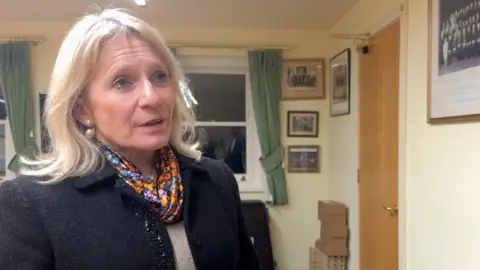 BBC Elaine Millar is wearing a black blazer, a floral scarf and she has blonde hair. She is speaking in a meeting room in St John's parish hall 