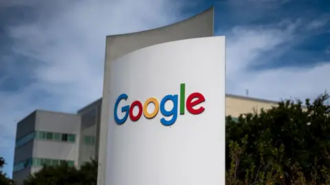 Bloomberg via Getty Images Google's multi-coloured logo is seen on a white sign in outside their headquarters, a grey and beige building with trees in the foreground and wispy clouds against a blue sky, in Mountain View, California in 2024