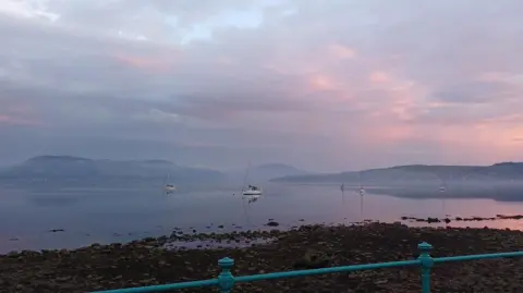 Elaine Sheard A calm shoreline scene with sailboats anchored on still water under a dramatic sky tinged with pink and purple hues. Mist hangs over the distant hills, while the water reflects the soft evening colours.