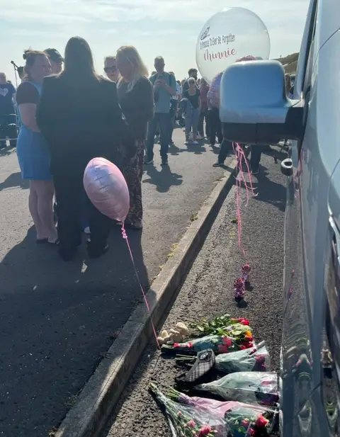 Flowers and a balloon beside a grey private ambulance with people in the background