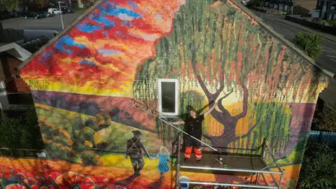 Dale Baxter/BBC Nikita stands on scaffolding in front of the mural holding a roller with her right hand. She has her arms outstretched and is smiling. She is wearing glasses, fluorescent orange work trousers and a black t-shirt. The mural: in the foreground we can see red poppies with fields stretching out behind them as the sun sets. A soldier walks away holding hands with his daughter. He is dressed in a military uniform and his daughter has blonde hair and a light blue dress. Above them there is a large willow tree.