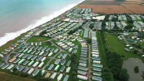 Parkdean An aerial view of Skipsea Sands holiday park in East Yorkshire, with dozens of colourful caravans arranged in rows close to a beach and the sea. To the right is a large green space and a lake.