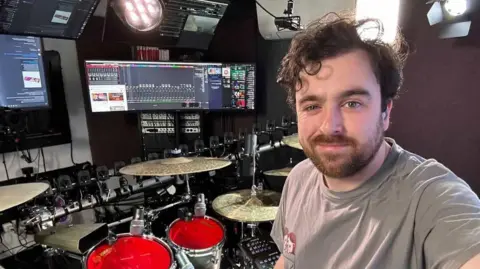 Greg Storey, a man with curly brown hair and a beard. He is in a recording studio with drums and cymbals in the background as well as screens and lights. He is wearing a grey t-shirt.