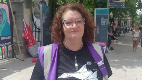 Helen Crickard standing at the Belfast protest, looking directly at the camera. She has shoulder-length curly brown hair and glasses. She is wearing a black t-shirt under a purple and grey gillet.