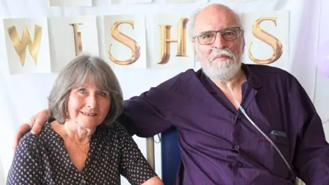A woman with shoulder-length grey hair in a spotted dress sitting down next to a bald elderly man with a white beard who is wearing a purple and blue shirt and has a nose tube in.