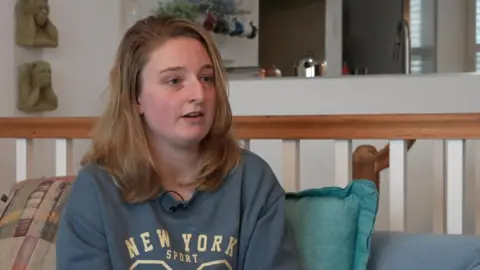 Emily Hotchkiss is sitting on a sofa talking. She has shoulder-length blonde hair and blue eyes. She is wearing a blue hoodie with the words: NEW YORK SPORT.
Behind her are cushions and a wooden bannister. There are two 'see no evil, speak no evil' sculptures hanging on the wall and a kitchen beyond.