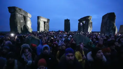 Andrew Matthews/PA Wire People gathered around the stones of Stonehenge under a dark sky - many holding smartphones and in woolly hats