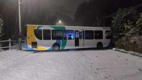 Chris Carnell A bus is blocking a road. The road is covered in snow.