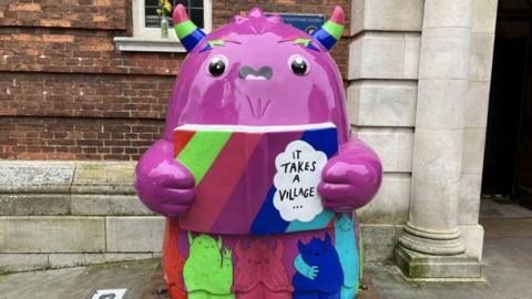 Pink Snook sculpture holding a book that says "it takes a village"