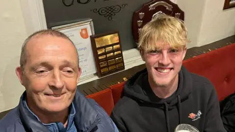 FAMILY HANDOUT Dave on theleft he has a bright smile and short grey hair, wearing a navy jacket, he sits next to his son Oli, who has blonde hair and a big grin.