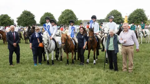 Five riders on horseback wearing rosettes sit for a photo in the middle of a field with trees and a number of other people standing nearby.