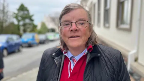 A man with grey and black shoulder length hair. He's wearing thin rimmed black glasses. He's wearing a blue Barbour jacket and a red jumper underneath.