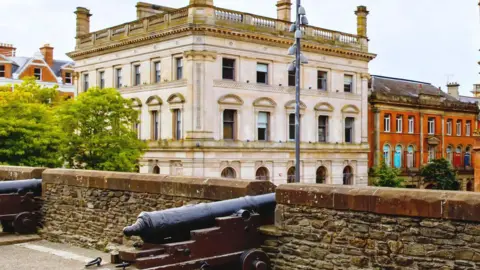 The former Danske Bank building is a large grey building. It is viewed from Derry's historic walls were cannons are seen up against the city walls. 