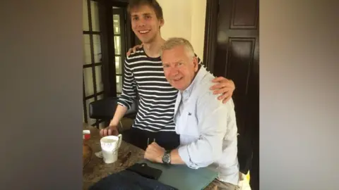 David Smith Two men are sitting in a home's kitchen at a granite countertop. The man on the left is wearing a long‑sleeved shirt with horizontal dark and light stripes. One arm is draped around the shoulders of the other person who is seated beside the countertop on the right side of the image. This person is wearing a light-colored button‑down shirt with sleeves rolled up.