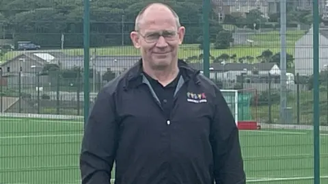 Karl Adamson Karl Adamson has short grey hair, he wears glasses and a black coat with the Orkney Island Games logo. He stands close to a green football pitch.